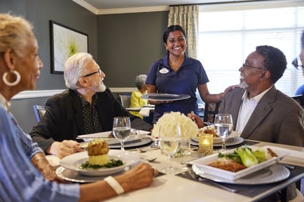 A smiling staff member serves a table of diners enjoying plated meals together, reflecting the resort-style amenities and warm hospitality of senior living in Delray Beach, FL.
