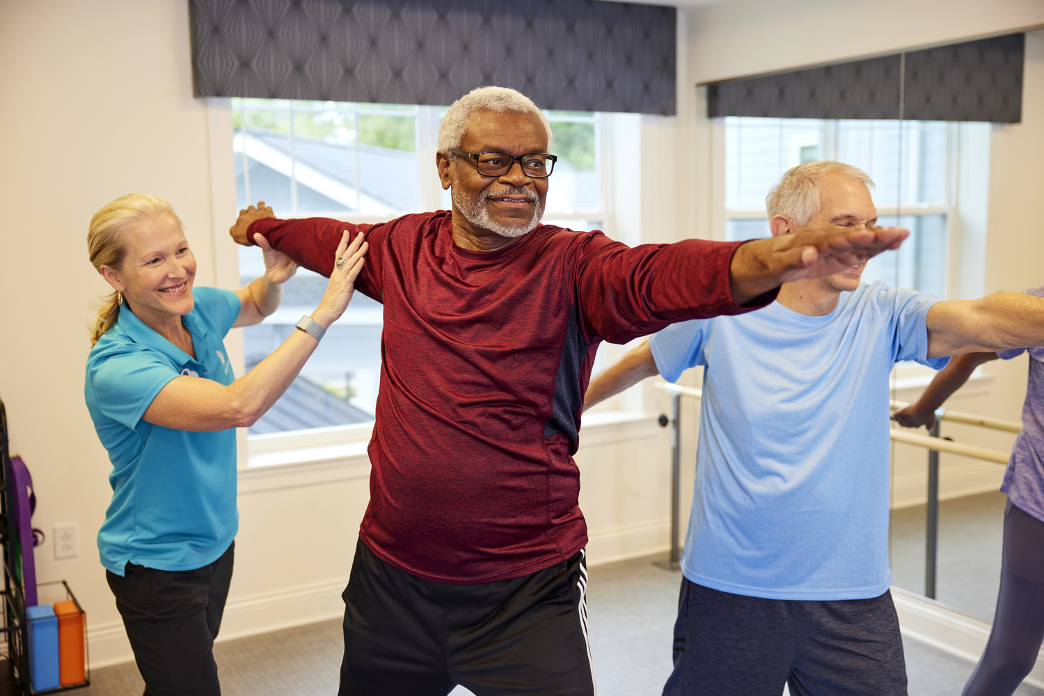 A staff member in a blue polo guides two older adults through a stretching exercise with arms extended at a memory care community in Fort Myers, FL, in a bright, open fitness studio.