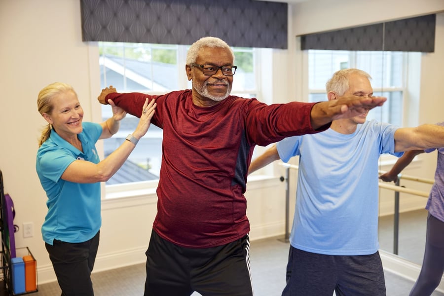 A staff member in a blue polo guides two older adults through a stretching exercise with arms extended at a memory care community in Fort Myers, FL, in a bright, open fitness studio.