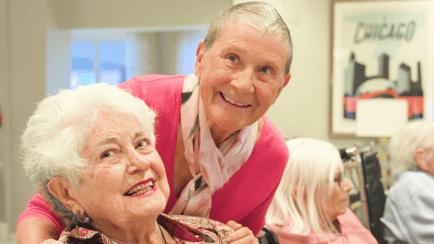 Two residents share a warm embrace and smile together during a group gathering, capturing the connection found in senior living in Glenview, IL.
