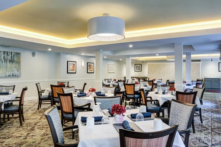 An elegantly appointed dining room with white-clothed tables, fresh floral centerpieces, and warm ambient lighting at Arbor Terrace Herndon, offering the refined senior living with dining and activities Herndon VA experience of resort-style independent living Herndon