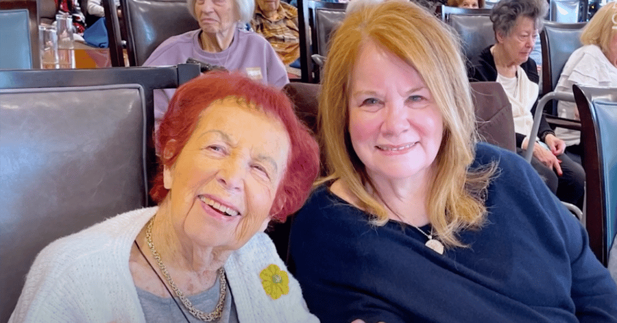 A resident with vibrant red hair smiles closely with a family member in a navy top, seated in a busy restaurant booth with other residents visible behind them. This reflects the active senior lifestyle and senior living in Highland Park, IL.