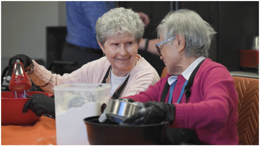 Two smiling residents bake together during a hands-on activity at Arbor Terrace Highland Park, embracing the active senior lifestyle in Highland Park, IL. Everyday moments like this reflect the warmth of senior living here.