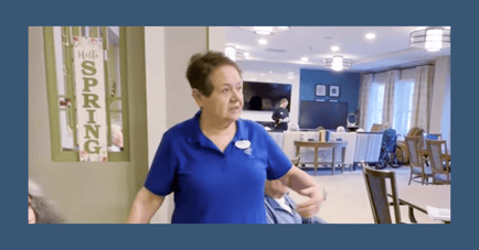 A staff member in a blue uniform chats in the sunny common area at Arbor Terrace Highland Park, decorated for spring. This personal touch reflects the senior living and senior care Highland Park, Illinois, families appreciate.