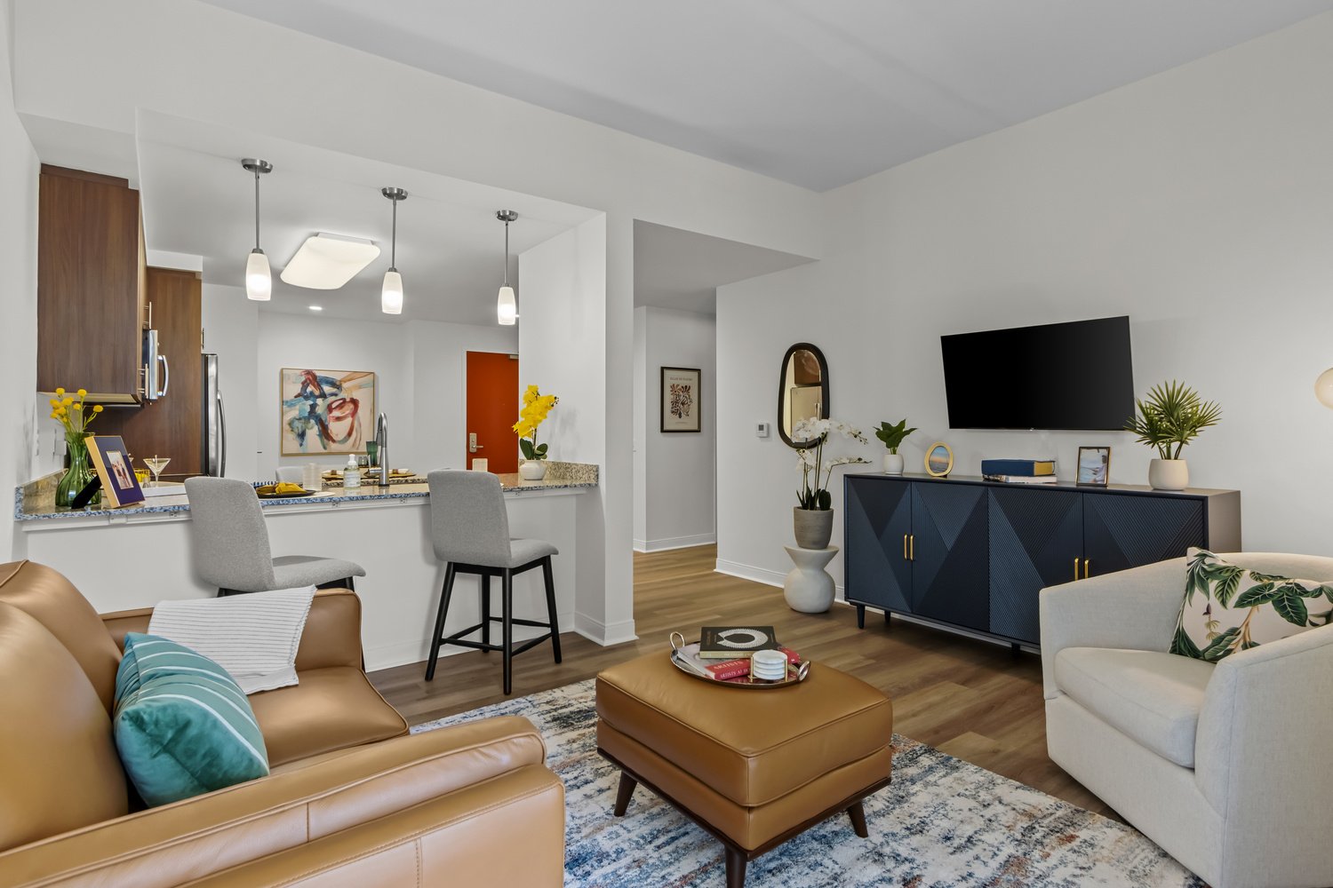 Lake Worth apartment living room with a carpet, a brown couch and matching ottoman, a white chair to the side. A tv stand and plants. A tv and art on the walls. A bar area with hanging lights. two stools looking into the kitchen