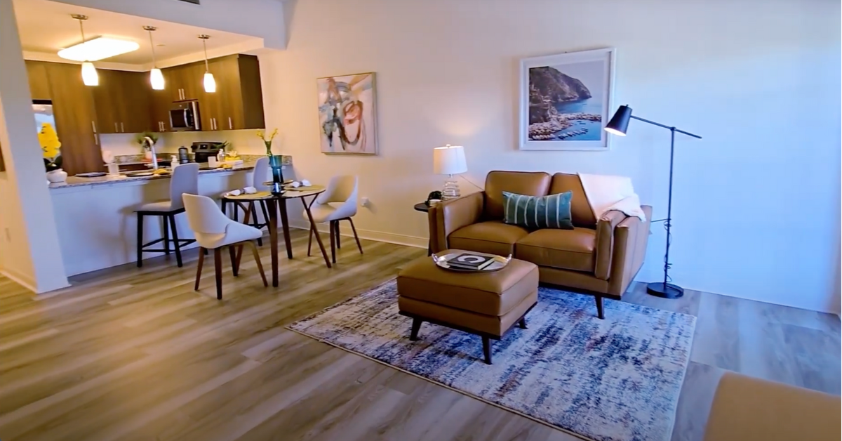 A living room area with a brown couch and brown ottoman. a rug on the floor and a blue pillow and photograph. Lamp to the right of the couch and a two person table to the left. Kitchen with wooden cabinets and bar area 