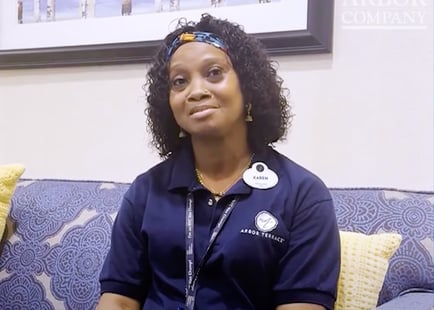 A staff member in a navy polo smiles warmly while seated on a patterned blue sofa with soft yellow pillows, reflecting the caring team behind senior care Mount Laurel New Jersey at one of the top senior living communities in Mount Laurel.