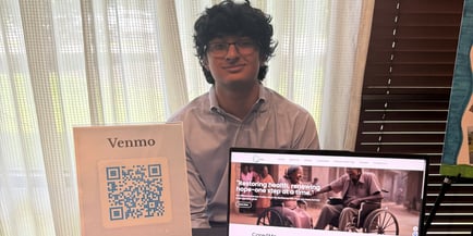 A young volunteer smiles while seated beside a Venmo donation sign and a laptop, showcasing the community spirit that supports personalized assisted living South Jersey at Arbor Terrace Mount Laurel.