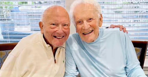 Two senior men beam with wide smiles while sitting close together indoors, capturing the joyful friendships and ageless spirit of life at Arbor Terrace Mountainside NJ's senior living community.