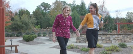 Two women walk hand in hand through a beautifully landscaped outdoor courtyard, smiling together at this assisted living community in Suwanee, GA