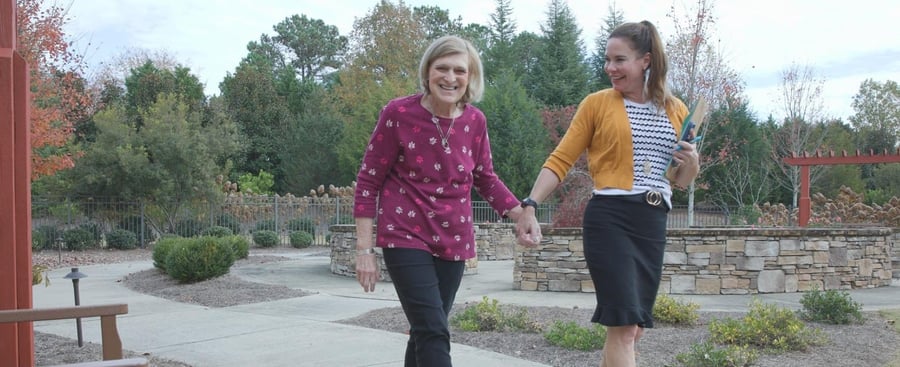 Two women walk hand in hand through a beautifully landscaped outdoor courtyard, smiling together at this assisted living community in Suwanee, GA