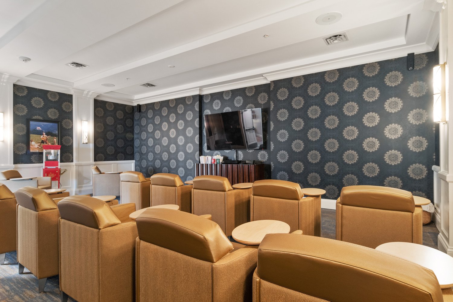 Arbor Terrace South Forsyth movie theatre with dark wallpapered walls with a tv on it. Tan chairs with little tables attached. Popcorn machine in the background