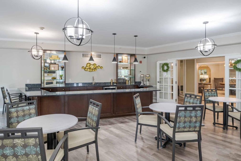 bistro area with wooden floors, white walls, pendant chandeliers, brown cabinets with white countertops, windows, white tables with three dining chairs each