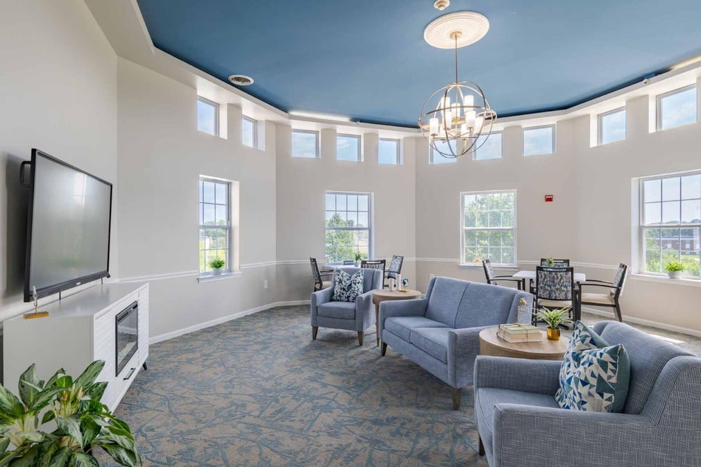 Circular common room with beige walls and abstract geometric carpeted floors. Big windows. Chandelier at the middle. Television on top of cabinet. Sofa and arm chairs in front of the television. Two tables with chairs. 