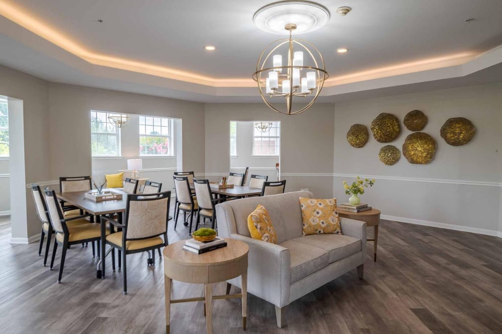 Common area with beige walls and wooden floors. Pendant chandelier with backlit ceiling lights. Artwork mounted on the wall. Two dining tables with chairs, loveseat with two coffee tables at each side. 