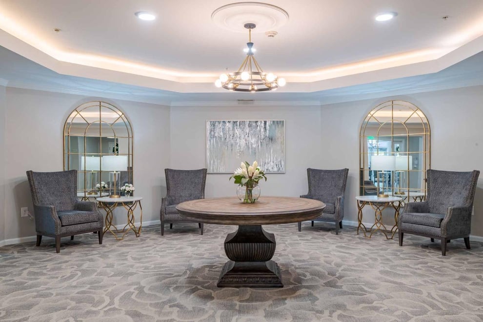 Common room with white walls and grey abstract carpeted floors. Round table at the middle with a flower on top and a circular backlit ceiling light with a chandelier above. Arm chairs against the walls. Big windows on the wall. 