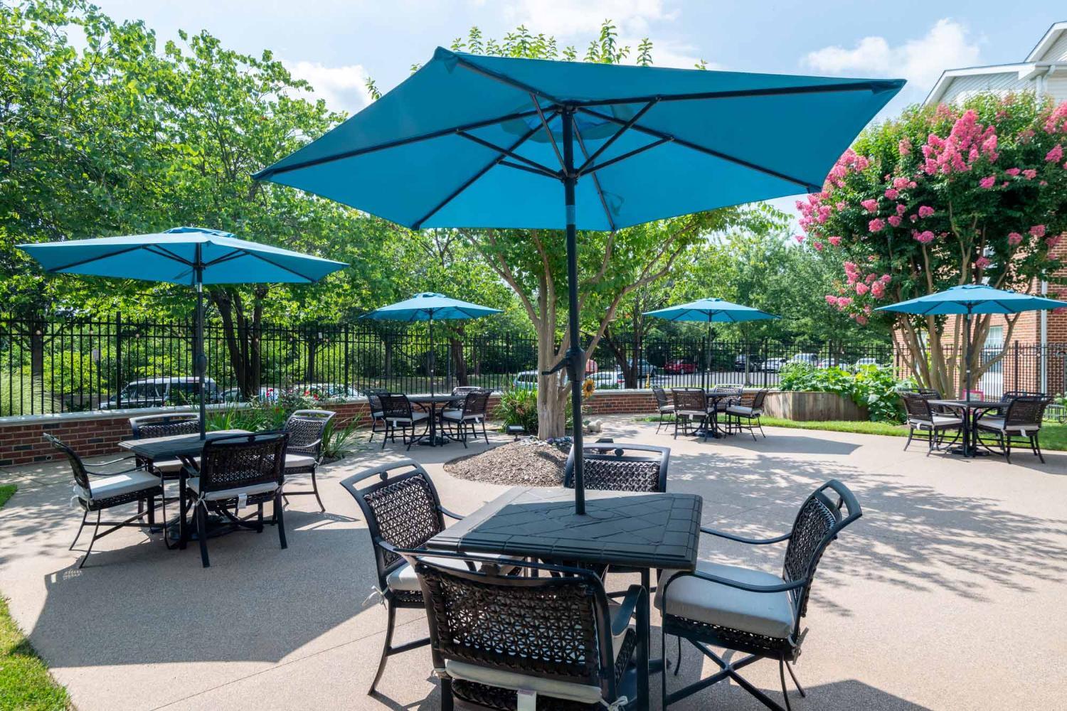 An outdoor sitting area, patio with tables and umbrellas and chairs, concrete floor, surrounded by trees and flowers