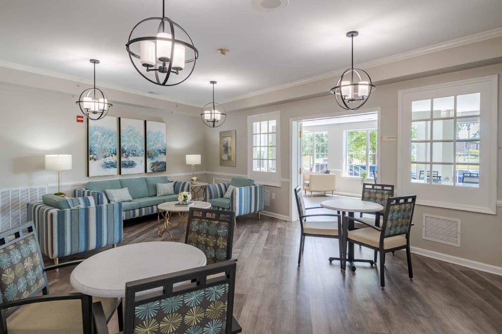 Dining area with beige walls and wooden floors. A sitting area with a blue sofa and matching arm chairs. Side tables on each sofa with lamps on top. Two round dining tables with chairs and pendant chandeliers above. Big windows and glass doors at one side. 