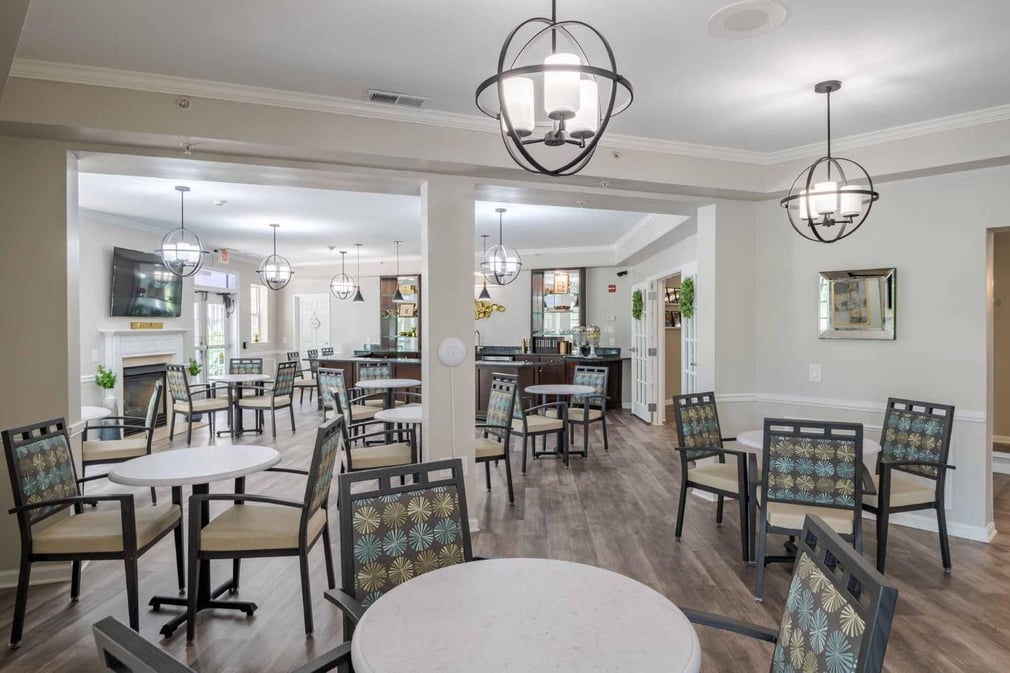Dining room with white walls and wooden floors. Multiple tables with chairs and pendant chandelier