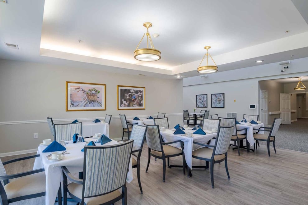 dining room with white walls, wooden floors, backlit ceiling lights. four paintings on the walls, multiple dining tables with table covers, padded dining chairs