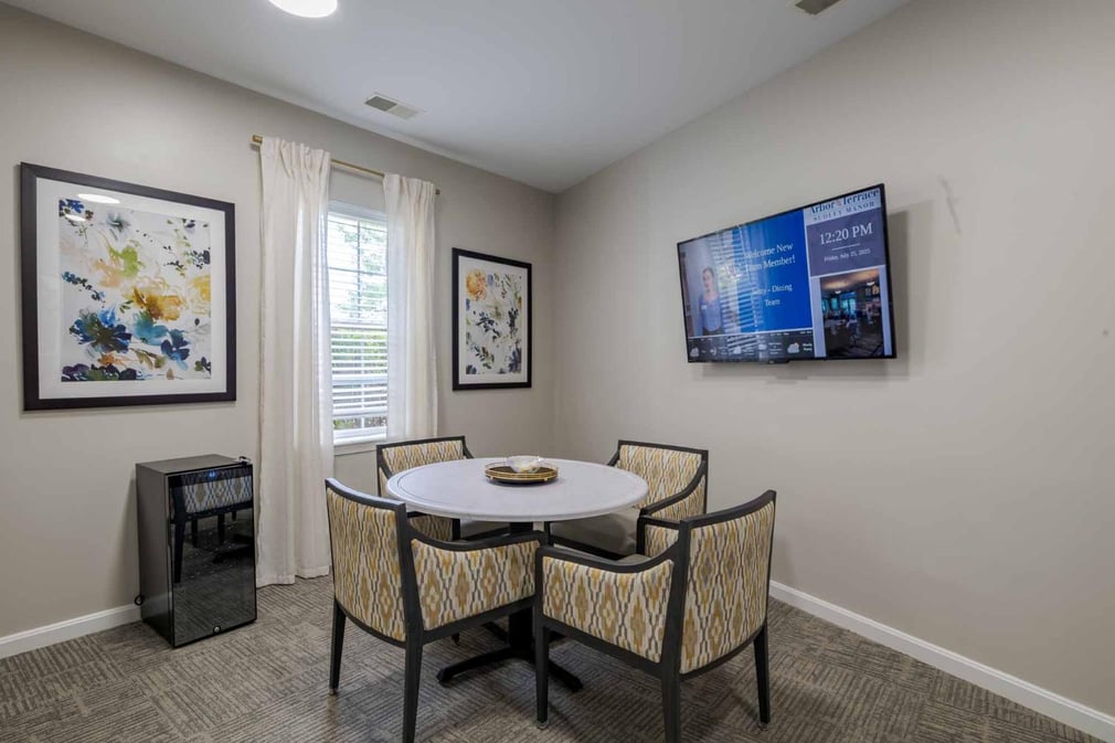 Entertainment area with wooden floors and beige walls. Television mounted on the wall, two paintings, a table with chairs. 