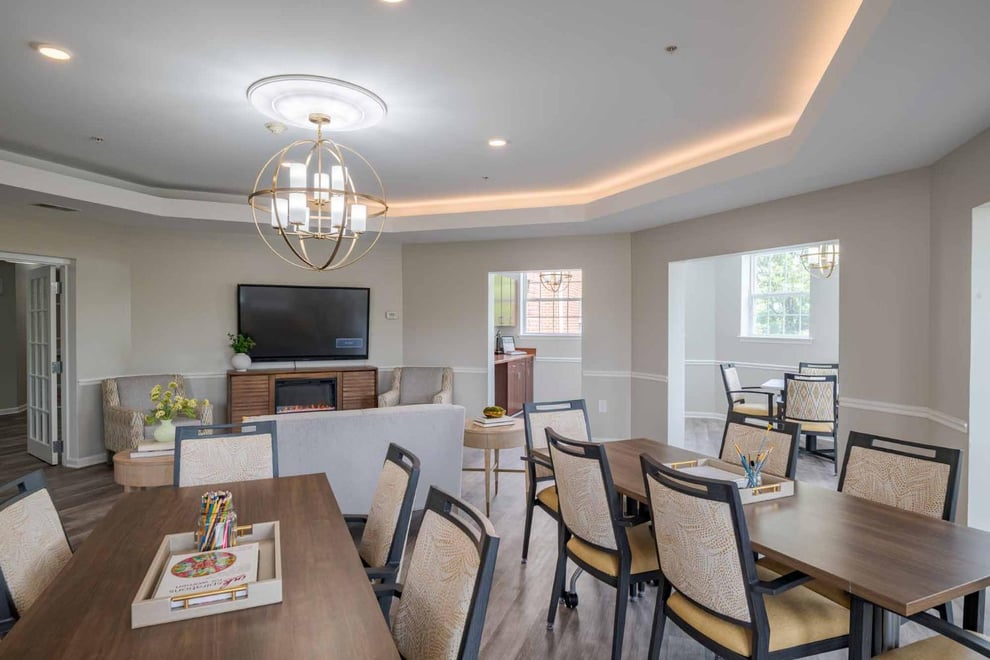 Event space with with white walls, wooden floors, back lit ceiling lights, chandelier. A fire place with a mounted TV on top, multiple tables and chairs, white sofa at the middle