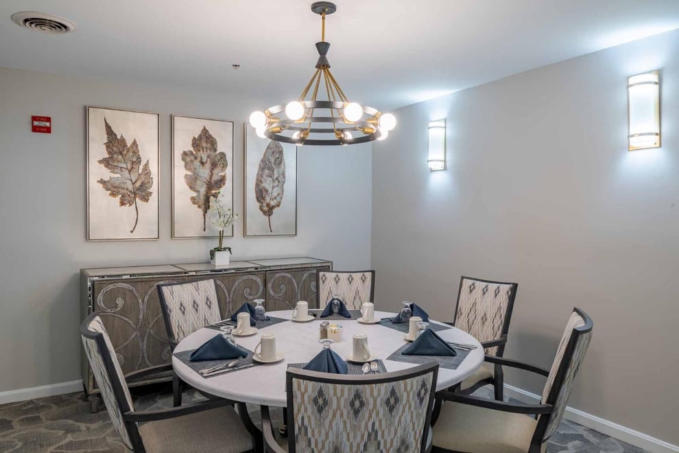 Dining room with white walls, grey abstract carpet, leaf artwork mounted on the wall, above a brown console table. six-seater round table with white padded chairs under round chandelier. Table set with utensils, mugs, and blue napkins