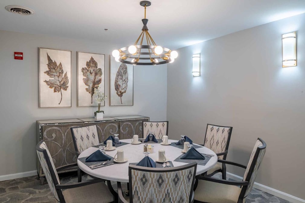 Dining room with white walls, grey abstract carpet, leaf artwork mounted on the wall, above a brown console table. six-seater round table with white padded chairs under round chandelier. Table set with utensils, mugs, and blue napkins