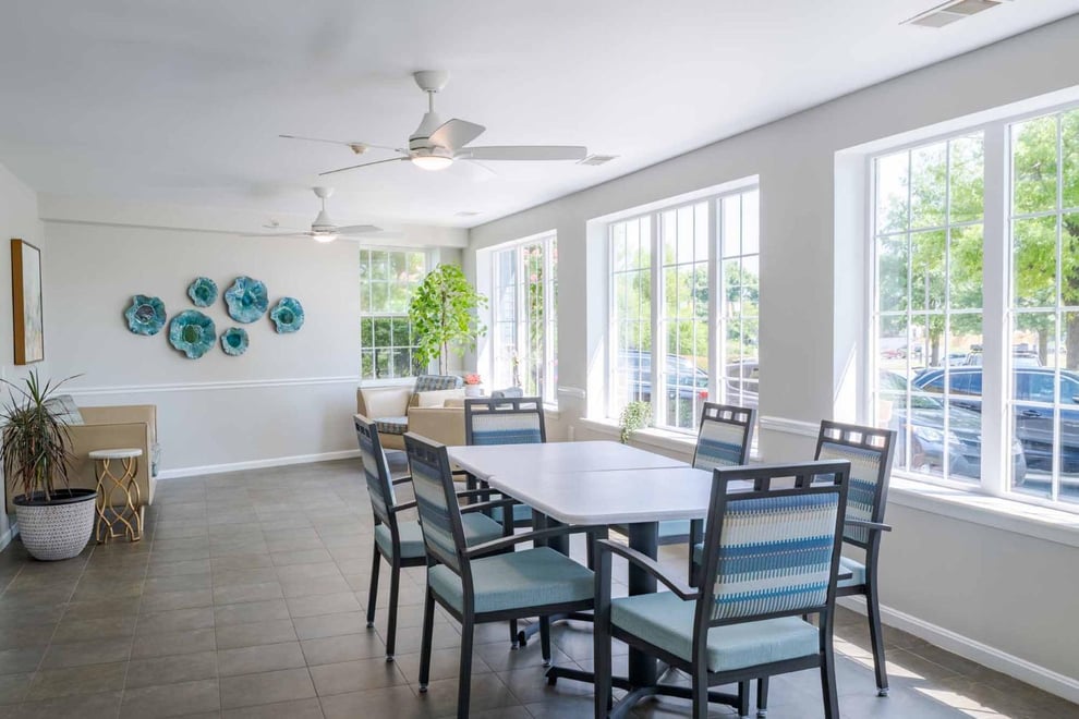dining area with white walls, wooden floors, ceiling fan, wide windows. Plants at the corner, art work on the farthest wall, six-seater dining table with dining chairs