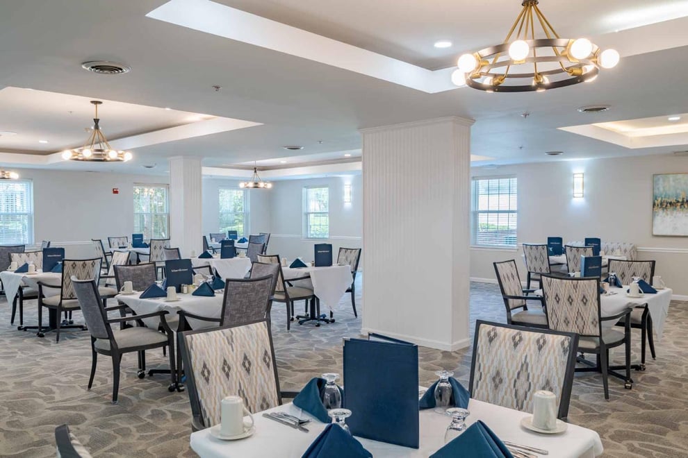 dining room with white walls, ivory carpet, wide windows, chandeliers, padded dining chairs with dining tables covered with white table cloth and utensils