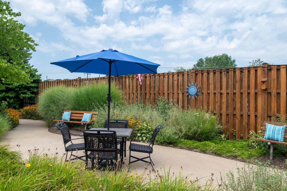 fenced outdoor area with greenery, table with umbrella, lawn chairs, and a bench