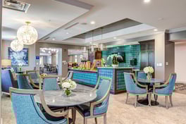Dining room area with blue chairs around circular tables with white flowers on top. blue tiled bar area in the back