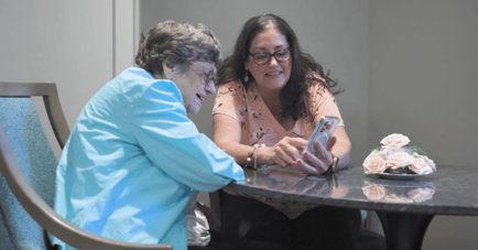 A resident and a visitor share a warm moment looking at a smartphone together at a table, reflecting the meaningful connections at our senior living community in Gambrills, MD.