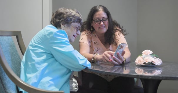 A resident and a visitor share a warm moment looking at a smartphone together at a table, reflecting the meaningful connections at our senior living community in Gambrills, MD.