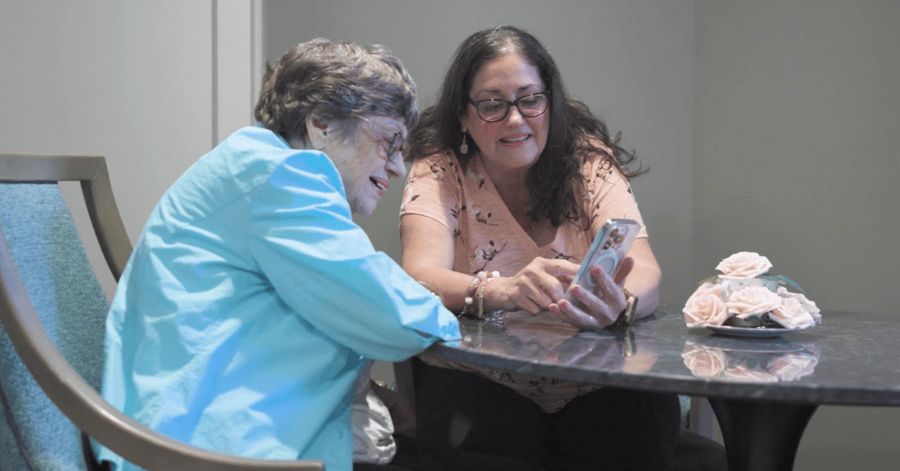 A resident and a visitor share a warm moment looking at a smartphone together at a table, reflecting the meaningful connections at our senior living community in Gambrills, MD.