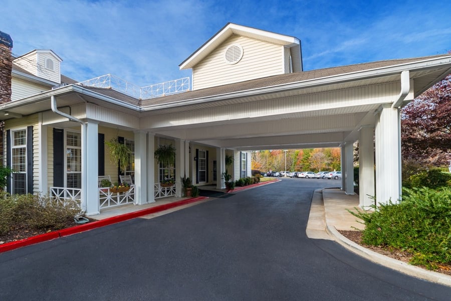 The welcoming front entrance of Arbor Terrace Athens, an assisted living community in Athens, GA, featuring a covered drop-off area, a cozy front porch with seating, and colorful autumn foliage surrounding the building.