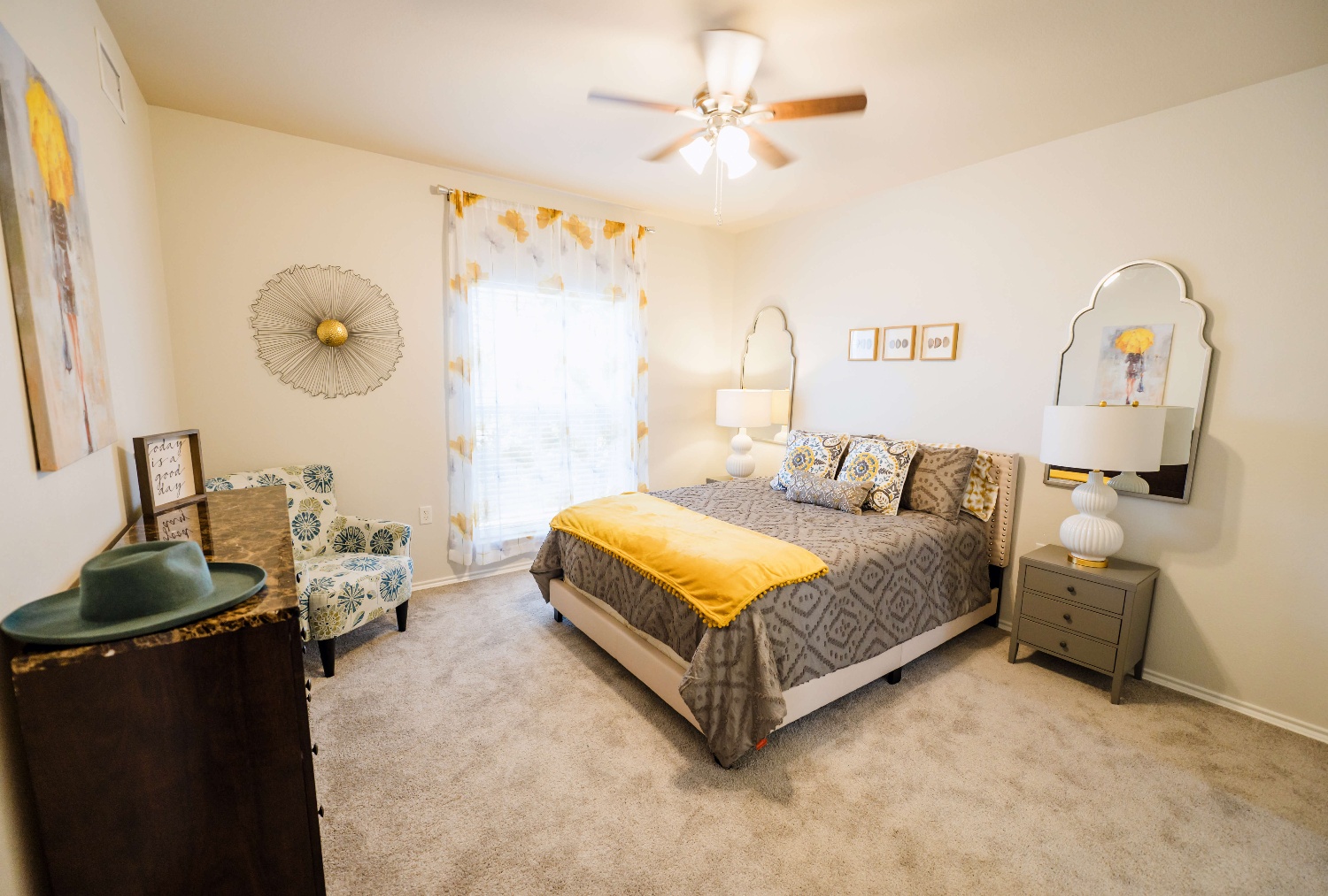 Carpeted bedroom with a large bed in the center with a brown and grey duvet and yellow blanket. two night stands on each side and mirror above. A brown dresser with a green hat sitting on it. artwork and decor along. the walls and a window. 