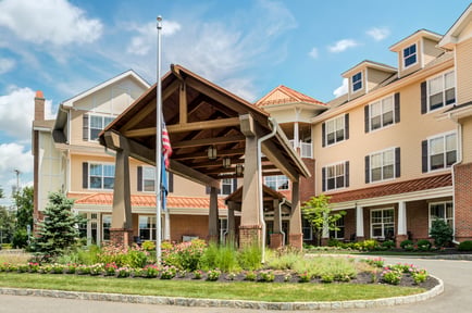 The welcoming exterior of Arbor Terrace Morris Plains features a covered porte-cochère, lush landscaping, and warm brick and wood accents, showcasing a beautiful assisted living community in Morris Plains, NJ.