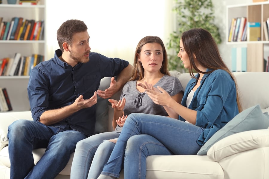 Three young adults sitting on a couch having an intense, emotional conversation, with a man and woman gesturing with their hands while another woman looks on with a concerned expression, in a living room with bookshelves in the background.