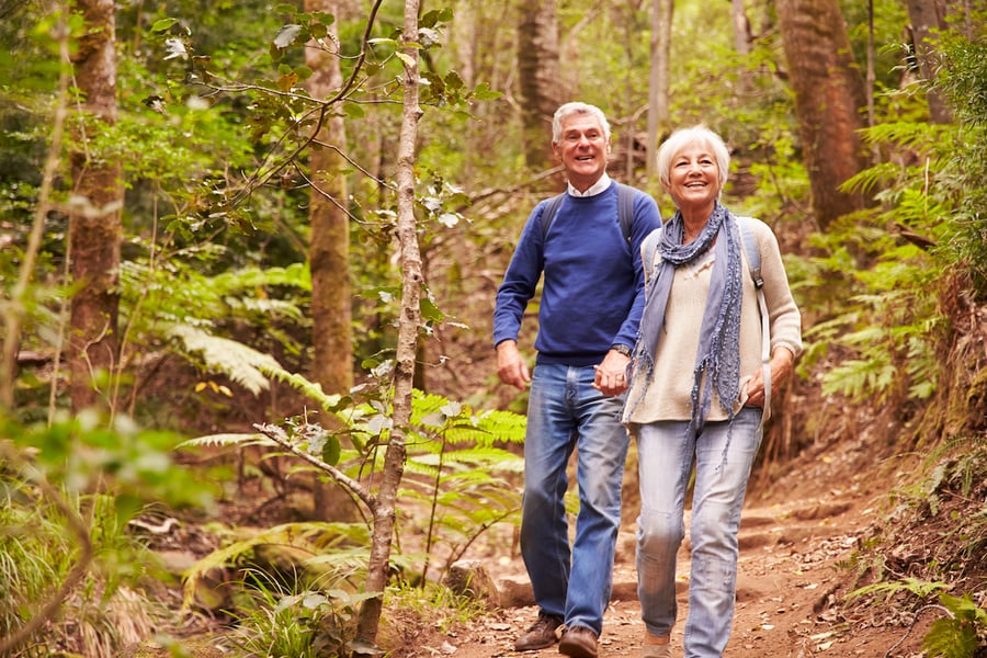 Smiling older couple walking side by side on a dirt trail through a lush, green forest with ferns and tall trees. The man wears a blue sweater and jeans, and the woman wears a cream sweater, a light blue scarf and jeans. Both carry backpacks.
