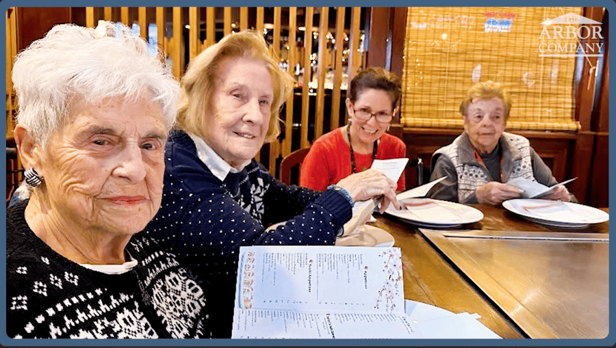 Residents and a staff member gather around a dining table with activity sheets at Arbor Terrace Morris Plains, highlighting the engaging, brain health-focused senior living programs available in Morris Plains, NJ.