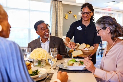 A dining staff member wearing a name badge warmly serves bread to laughing residents at a beautifully set table with fresh flowers, capturing the vibrant resort-style senior living experience in Woodbridge, VA.