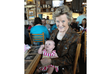 A smiling older adult holds a young baby at a restaurant table surrounded by other diners — a warm resident spotlight moment from Arbor Terrace Lakeway senior care community TX.