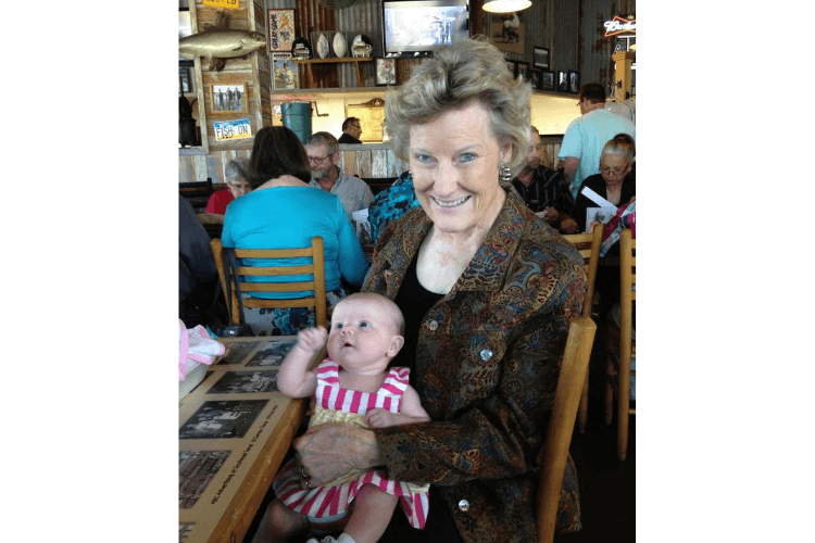 A smiling older adult holds a young baby at a restaurant table surrounded by other diners — a warm resident spotlight moment from Arbor Terrace Lakeway senior care community TX.