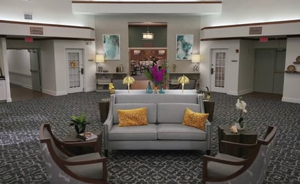 A spacious, welcoming common area at assisted living in Fort Myers, FL, with a gray sofa accented by gold pillows, dark wood armchairs, patterned flooring, warm lamps, and colorful artwork on the walls.