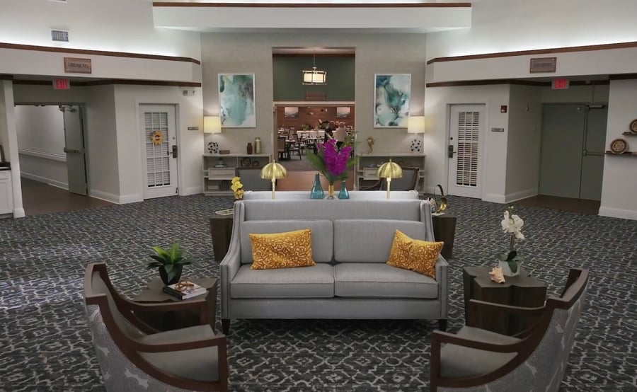 A spacious, welcoming common area at assisted living in Fort Myers, FL, with a gray sofa accented by gold pillows, dark wood armchairs, patterned flooring, warm lamps, and colorful artwork on the walls.