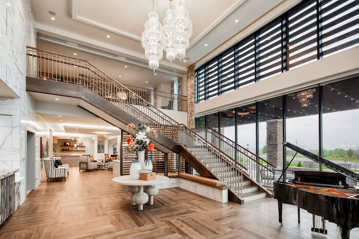 Arbor Terrace Fulton lobby area with wooden floors, a grand piano, and a round table in the center with flowers sitting on top. a grand stair case in the back and a common area. tall windows and high ceilings on and hanging crystal chandeliers.