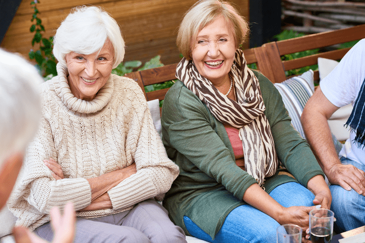 Two older adults smile warmly while seated outdoors on a wooden bench, enjoying friendly conversation at Arbor Terrace Herndon, an active senior living Herndon VA community offering vibrant independent senior living Herndon Virginia.