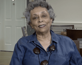 A woman smiles warmly while seated in a bright room, sharing her experience as part of the welcoming senior living community at Arbor Terrace Herndon in Herndon, VA.