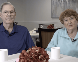 Two people sit together at a dining table with a floral centerpiece, smiling warmly in a cozy setting at Arbor Terrace Herndon, a welcoming senior living community in Herndon, VA.